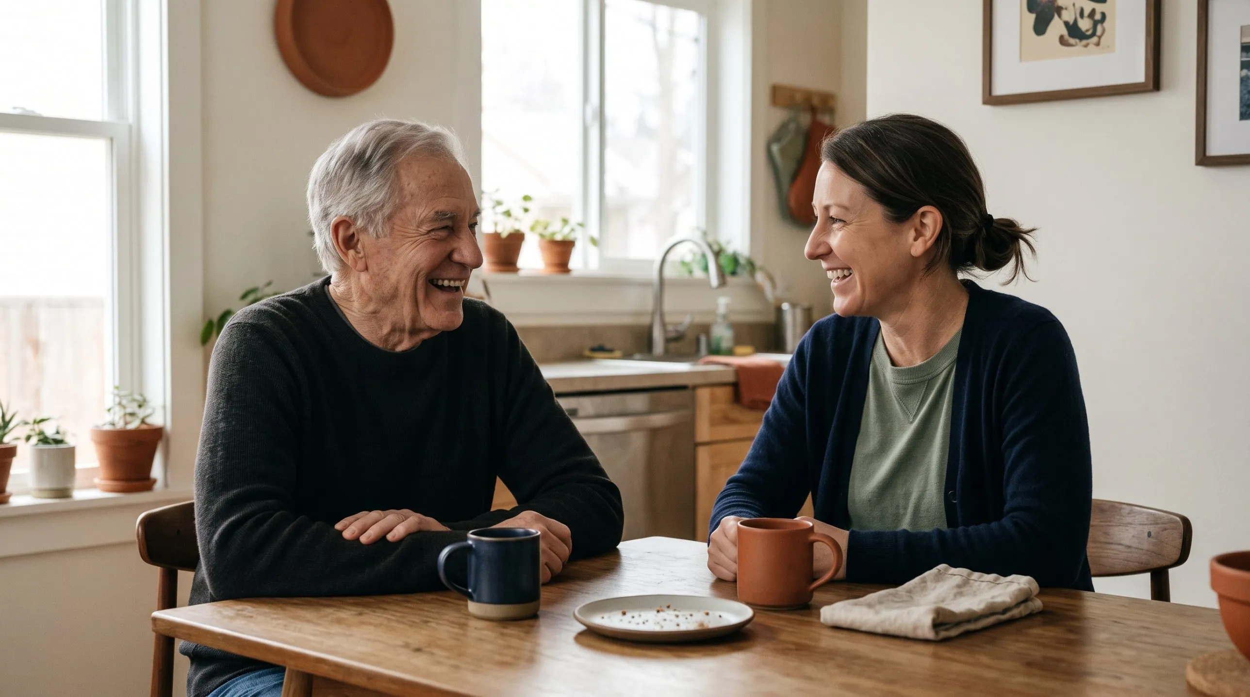 An older man and a younger woman laughing together over coffee at a kitchen table